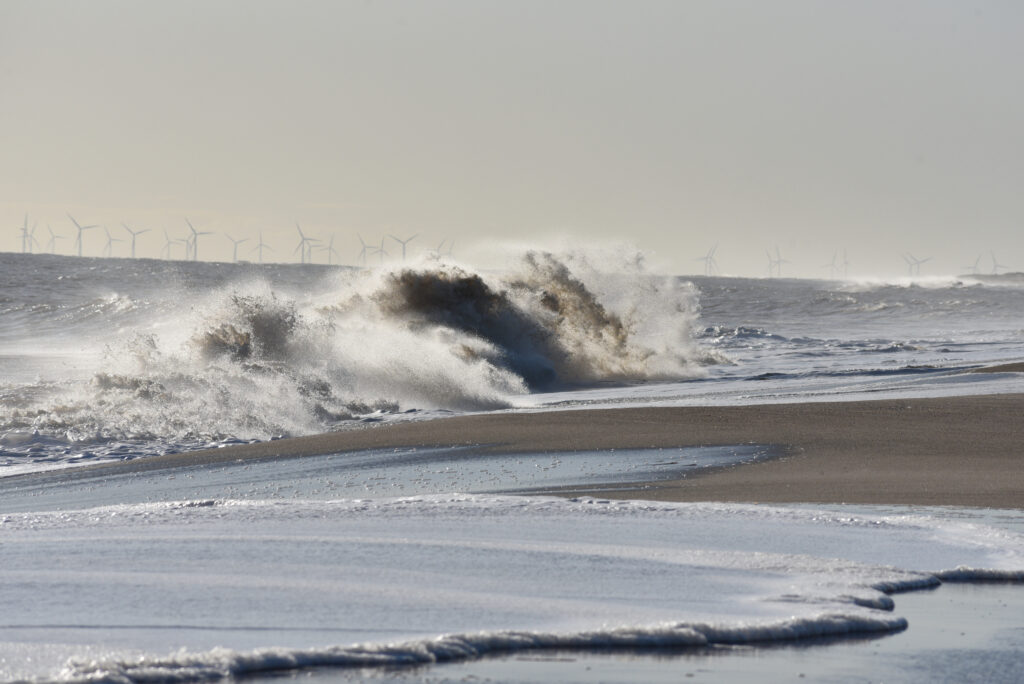 Waves crashing onto a sandy beach with offshore wind turbines on the horizon