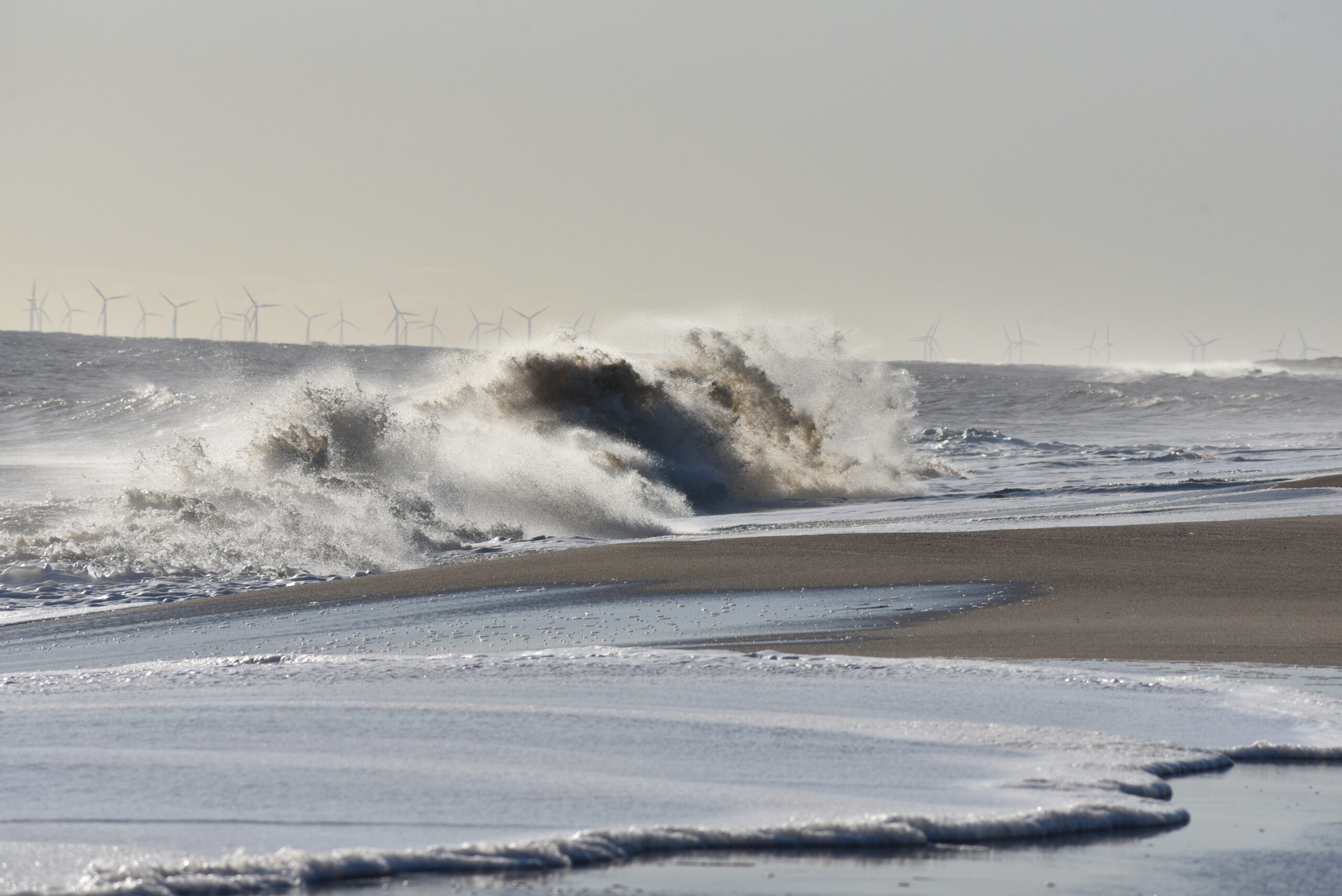 Waves crashing onto a sandy beach with offshore wind turbines on the horizon