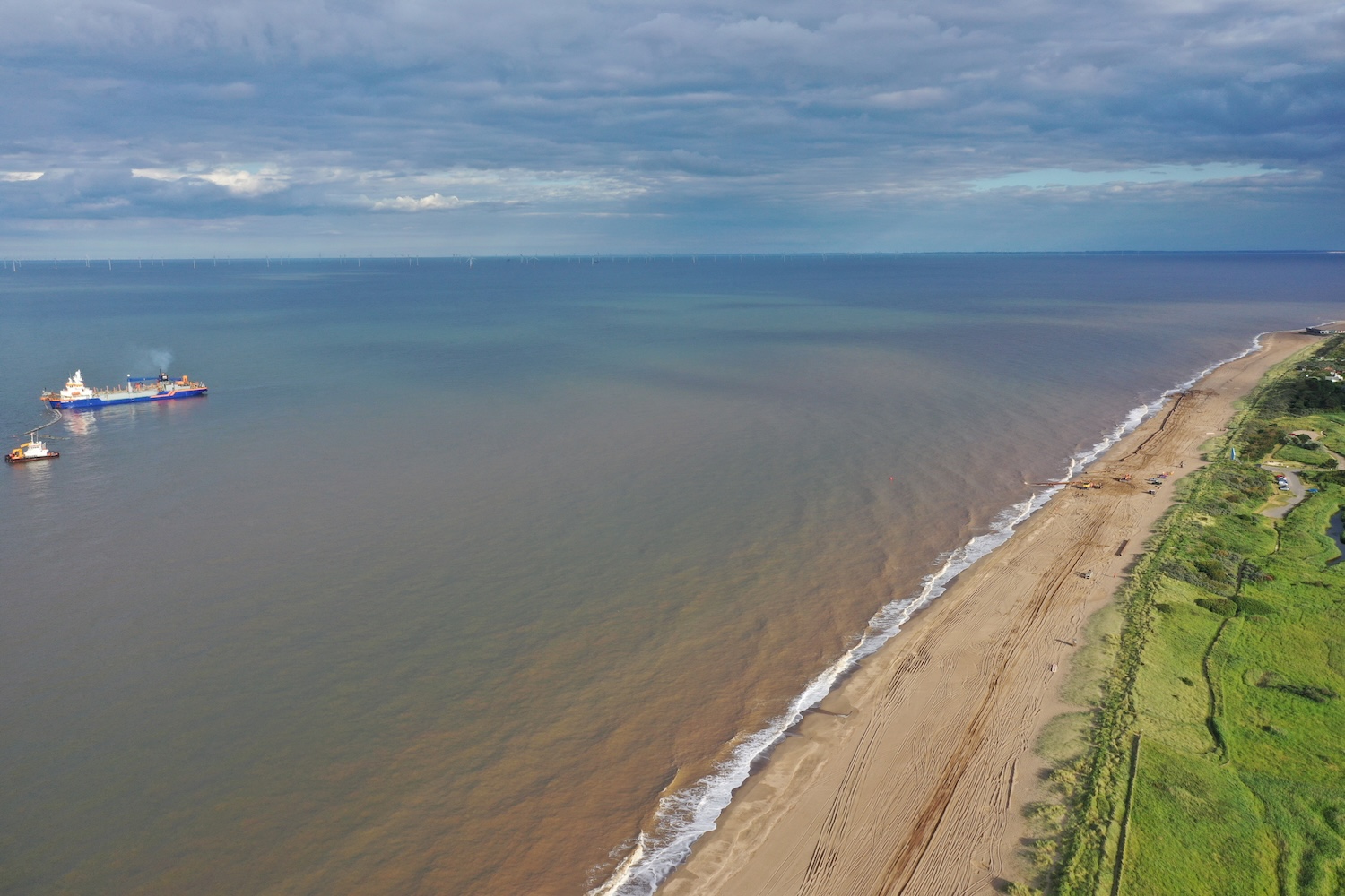 Aerial view of a sandy beach with grassy areas, gentle waves, and two ships in the water under a mostly cloudy sky.